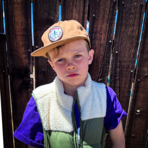 Boy wearing a tan cap with a circular patch, standing in front of a wooden fence.
