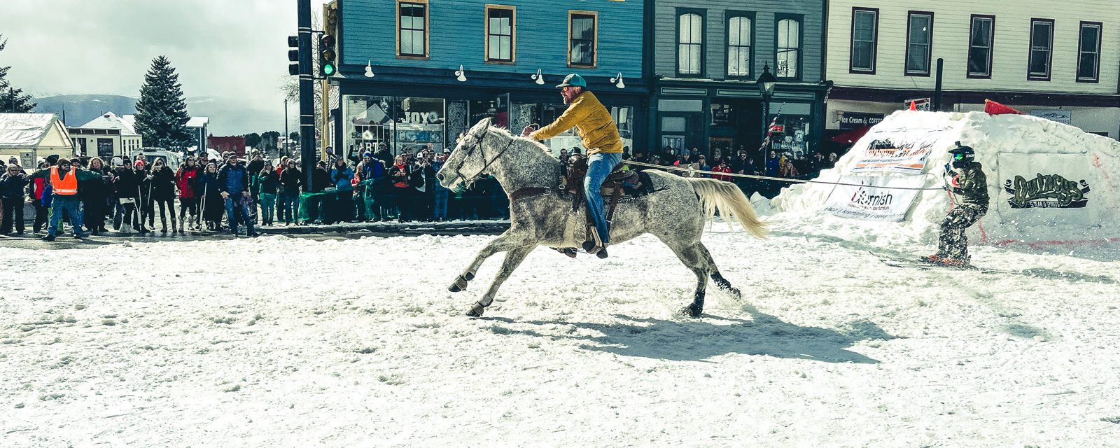 Leadville skijoring on Harrison Avenue: a rider on a gray horse tows a skier past the crowd and the historic storefronts